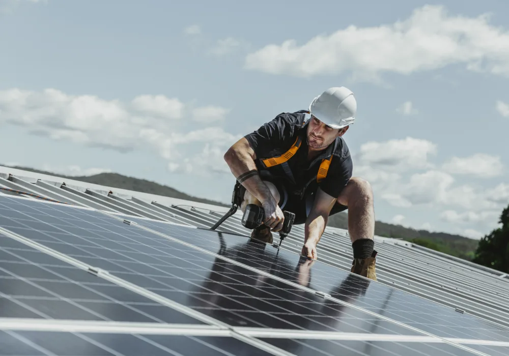 Ein Solarmonteur installiert Solarpanels auf einem Hausdach. Im Hintergrund ist der blaue Himmel mit einzelnen weissen Wolken zu sehen.