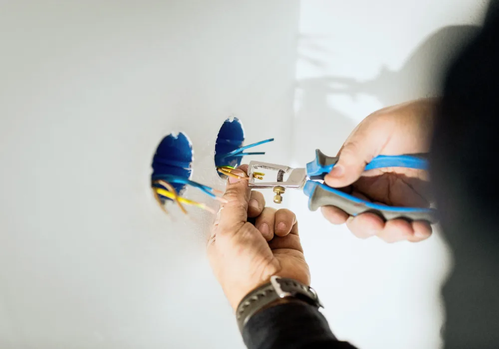 Ein Elektriker installiert eine Steckdose. Er hält eine Zange in der Hand, aus zwei Löchern in der Wand ragen Kabel, die der Elektriker gerade bearbeitet