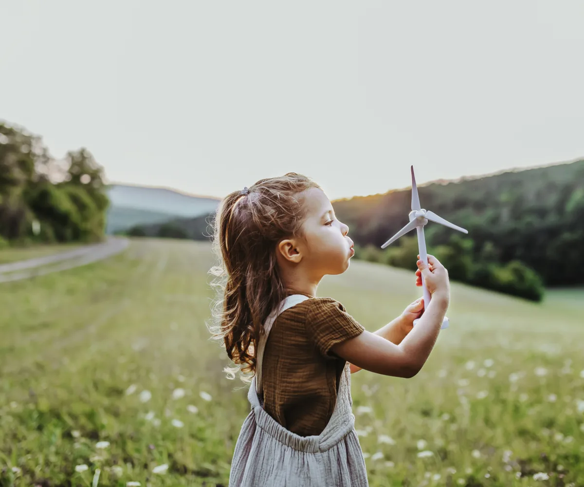 Ein junges Mädchen steht mitten in einer Blumenwiese und pustet statt einer Pusteblume eine kleine Spielzeug-Windkraftanlage