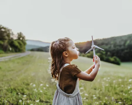 Ein junges Mädchen steht mitten in einer Blumenwiese und pustet statt einer Pusteblume eine kleine Spielzeug-Windkraftanlage