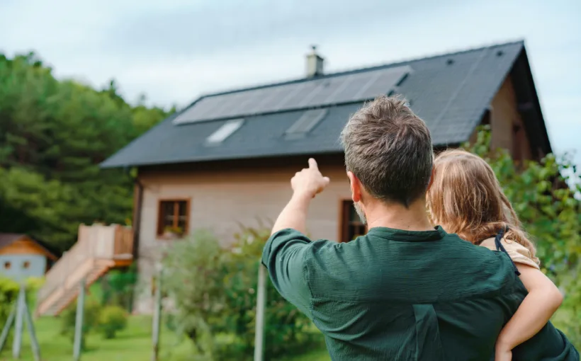 Ein Vater hält sein Kind auf dem Arm und zeigt ihm die Solarpanels auf ihrem Hausdach. Dabei handelt es sich um eine Eigenverbrauchsvariante