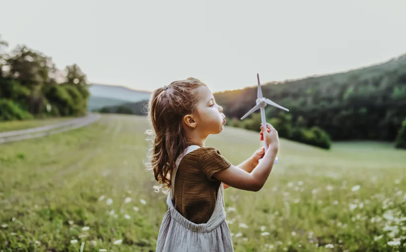 Ein junges Mädchen steht mitten in einer Blumenwiese und pustet statt einer Pusteblume eine kleine Spielzeug-Windkraftanlage