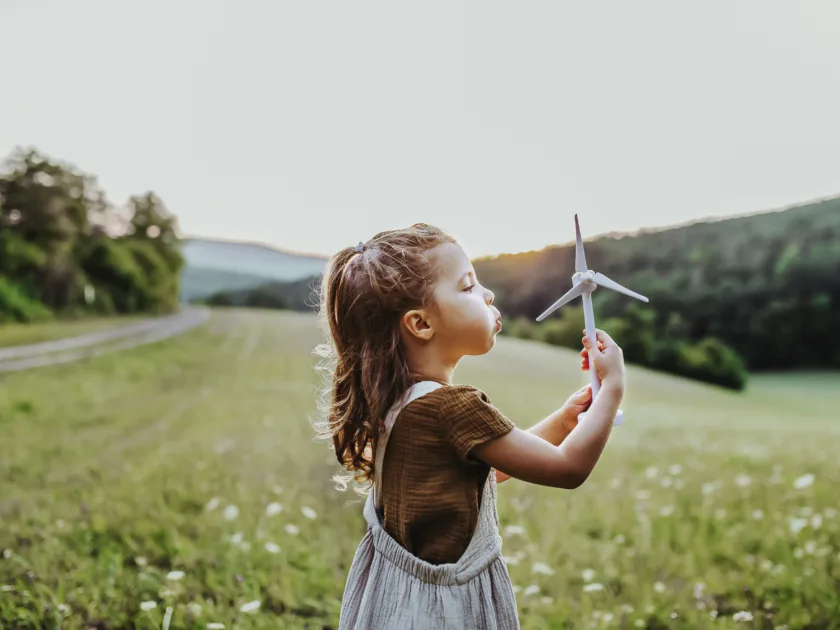 Ein junges Mädchen steht mitten in einer Blumenwiese und pustet statt einer Pusteblume eine kleine Spielzeug-Windkraftanlage