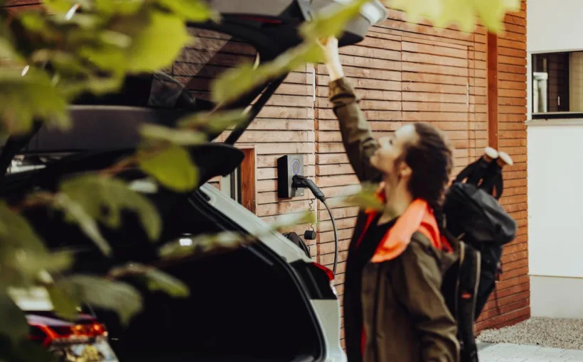 Eine Frau mit Wanderrucksack schliesst den Kofferraum ihres Elektroautos. Im Hintergrund ist die Ladestation zu sehen, an der das Auto angeschlossen ist.