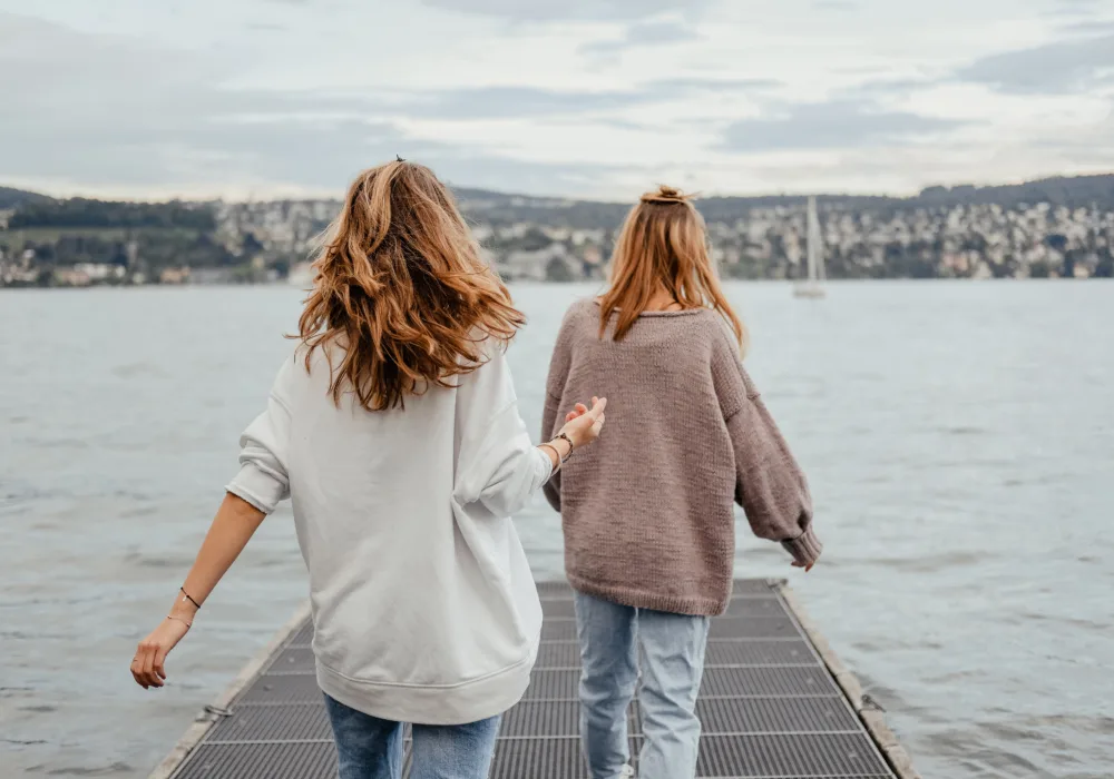 Zwei junge Frauen aus der Perspektive von hinten. Sie betreten in beschwingtem Gang einen Gittersteg, der auf den Vierwaldstättersee hinausführt. Im Hintergrund ist ein Segelboot und am Horizont das Ufer mit Teilen der Stadt Luzern zu sehen.