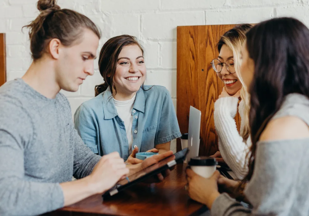 Vier junge Menschen stehen lachend an einem Tisch, eine Person bedient ein Tablet