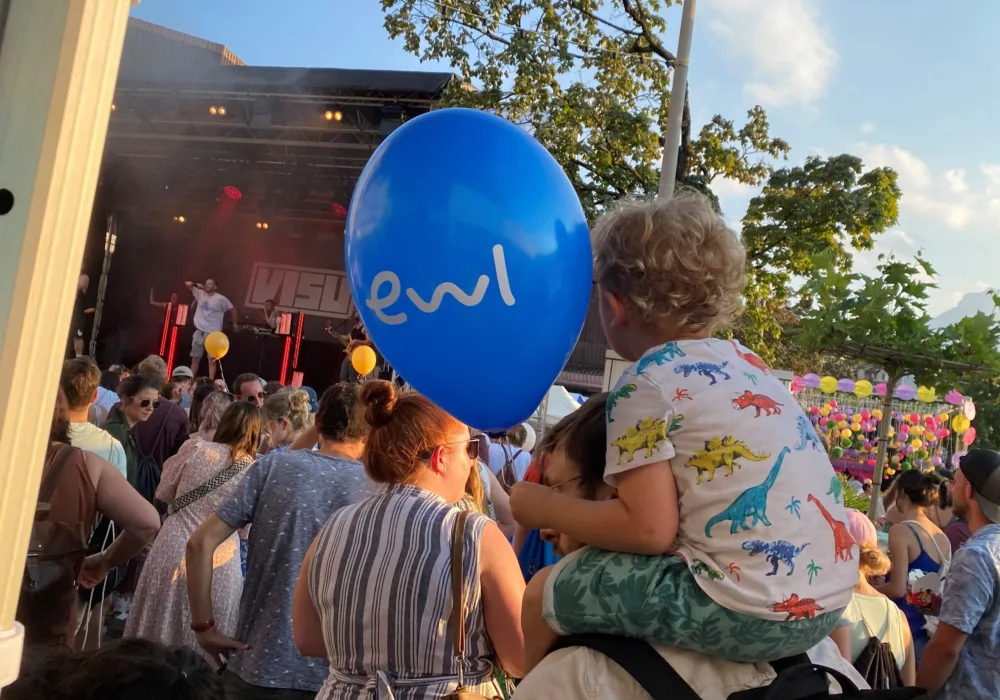 Das Foto zeigt einen von ewl gesponserten Live-Event. Im Hintergrund ist die Bühne sichtbar, auf der gerade ein Konzert aufgeführt wird. Die Perspektive zeigt von hinten auf die Bühne. Im Vordergrund ist das Publikum zu sehen. Gut sichtbar ein Vater, der ein Kleinkind auf der Schulter trägt. Das Kind hat einen blauen ewl-Luftballon in der Hand.