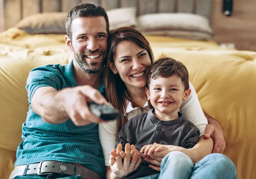 Vater, Mutter und Sohn sitzen im Wohnzimmer, der Vater richtet die Fernbedienung auf den Fernseher.