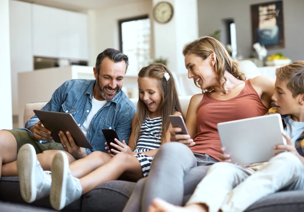 Eine Familie sitzt im Wohnzimmer auf dem Sofa. Vater, Tochter, Mutter, Sohn halten ein Tablet oder ein Smartphone in der Hand. Die Atmosphäre ist entspannt, alle lachen und sind vergnügt.