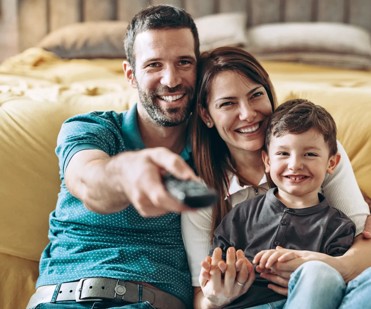 Vater, Mutter und Sohn sitzen im Wohnzimmer, der Vater richtet die Fernbedienung auf den Fernseher.