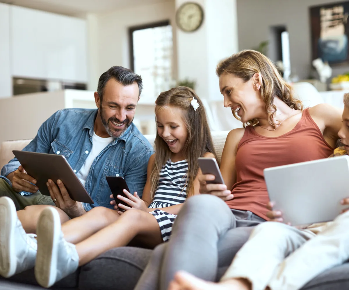 Eine Familie sitzt im Wohnzimmer auf dem Sofa. Vater, Tochter, Mutter, Sohn halten ein Tablet oder ein Smartphone in der Hand. Die Atmosphäre ist entspannt, alle lachen und sind vergnügt.