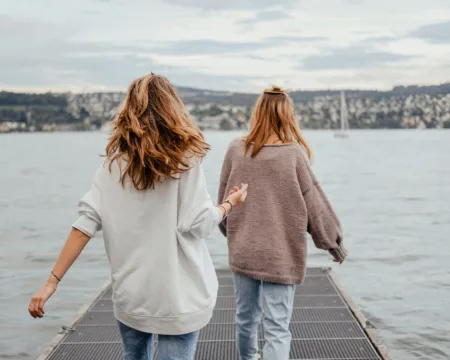 Zwei junge Frauen aus der Perspektive von hinten. Sie betreten in beschwingtem Gang einen Gittersteg, der auf den Vierwaldstättersee hinausführt. Im Hintergrund ist ein Segelboot und am Horizont das Ufer mit Teilen der Stadt Luzern zu sehen.