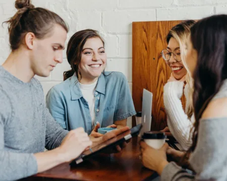 Vier junge Menschen stehen lachend an einem Tisch, eine Person bedient ein Tablet