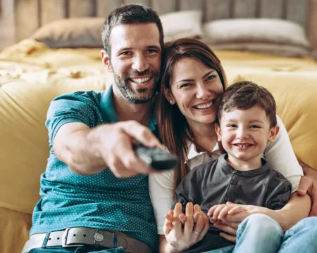 Vater, Mutter und Sohn sitzen im Wohnzimmer, der Vater richtet die Fernbedienung auf den Fernseher.