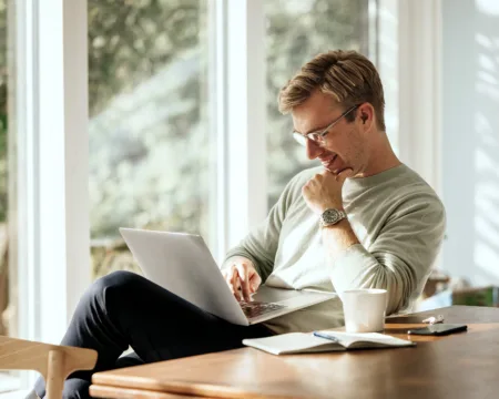 Ein junger Mann sitzt lachend am Tisch und bedient auf seinem Schoss einen Laptop