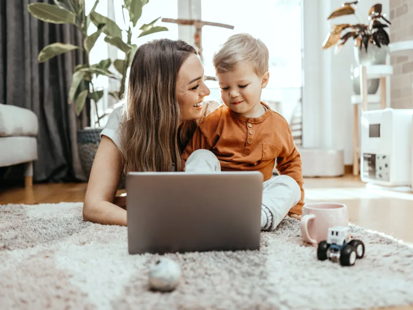 Eine Mutter sitzt mit ihrem Kind im Wohnzimmer auf dem Teppich am Boden, vor ihnen steht ein Laptop. Es sind weitere Details wie Spielzeug und Kaffeetasse sichtbar, im Hintergrund Wohnzimmermöbel