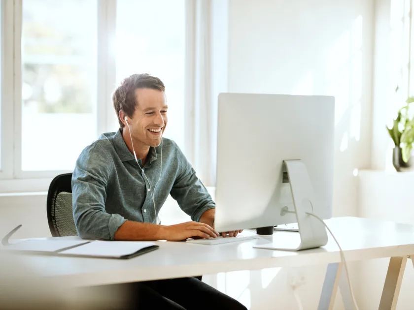 Junger Mann im Homeoffice. Er sitzt lachend am Bildschirm und trägt Kopfhörer