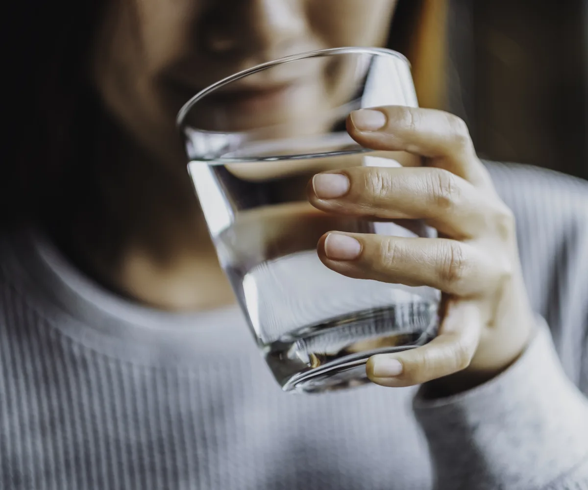 Eine Frau hält ein Glas mit Luzerner Trinkwasser in der Hand. Im Hintergrund sieht man ihr angedeutetes Lächeln. Sie führt das Glas zum Gesicht, um einen Schluck zu nehmen.