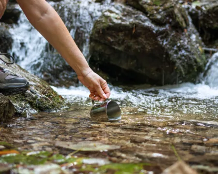 Eine Person in Wanderschuhen schöpft mit einem Trinkbecher Wasser direkt ab der Quelle eines Bergbachs