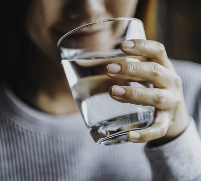 Eine Frau hält ein Glas mit Luzerner Trinkwasser in der Hand. Im Hintergrund sieht man ihr angedeutetes Lächeln. Sie führt das Glas zum Gesicht, um einen Schluck zu nehmen.