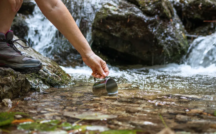 Eine Person in Wanderschuhen schöpft mit einem Trinkbecher Wasser direkt ab der Quelle eines Bergbachs