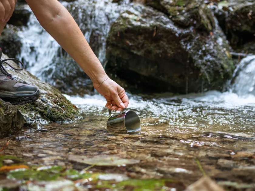 Eine Person in Wanderschuhen schöpft mit einem Trinkbecher Wasser direkt ab der Quelle eines Bergbachs