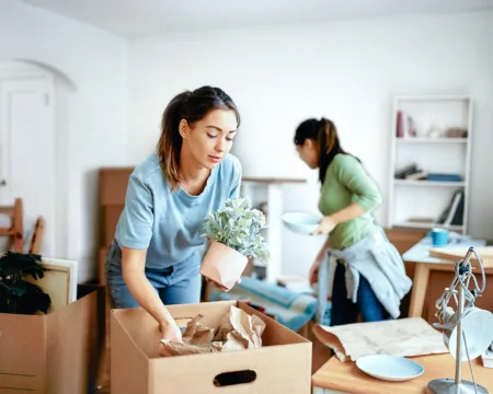 Zwei junge Frauen bereiten im Wohnzimmer ihren Umzug vor. Das Zimmer ist mit gepackten Kisten überfüllt. Teilweise stehen noch Möbel rum.