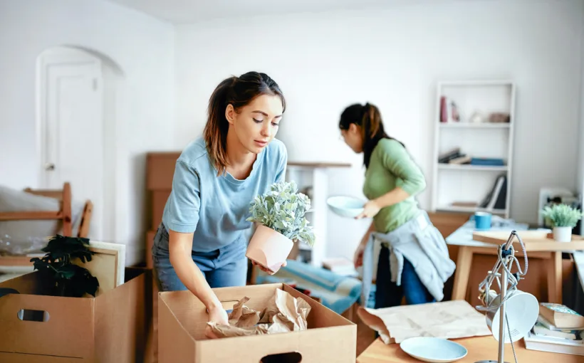 Zwei junge Frauen bereiten im Wohnzimmer ihren Umzug vor. Das Zimmer ist mit gepackten Kisten überfüllt. Teilweise stehen noch Möbel rum.