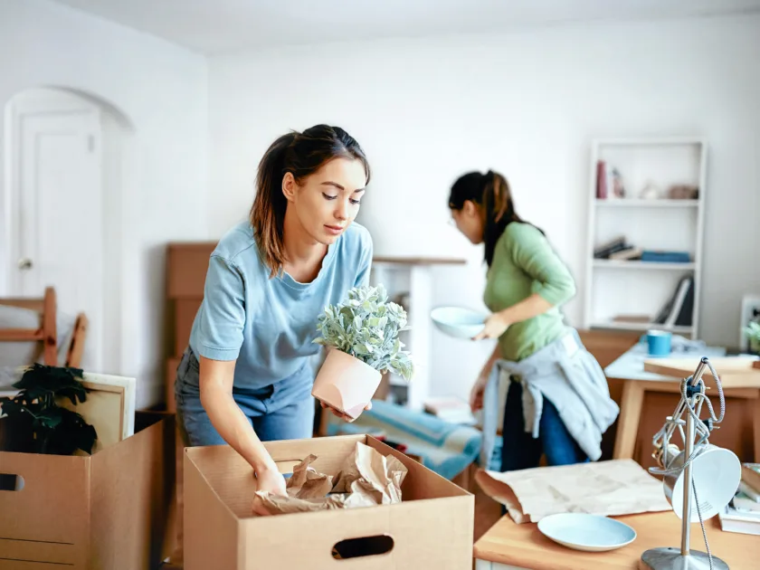 Zwei junge Frauen bereiten im Wohnzimmer ihren Umzug vor. Das Zimmer ist mit gepackten Kisten überfüllt. Teilweise stehen noch Möbel rum.