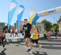 Ein Eventfoto des Luzerner Stadtlaufs am Standort Mühlenplatz. Im Vordergrund sind ein erwachsener und ein jugendlicher Teilnehmer zu sehen. Im Hintergrund sehen wir verschiedene Sponsoring-Elemente von ewl