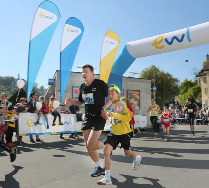 Ein Eventfoto des Luzerner Stadtlaufs am Standort Mühlenplatz. Im Vordergrund sind ein erwachsener und ein jugendlicher Teilnehmer zu sehen. Im Hintergrund sehen wir verschiedene Sponsoring-Elemente von ewl