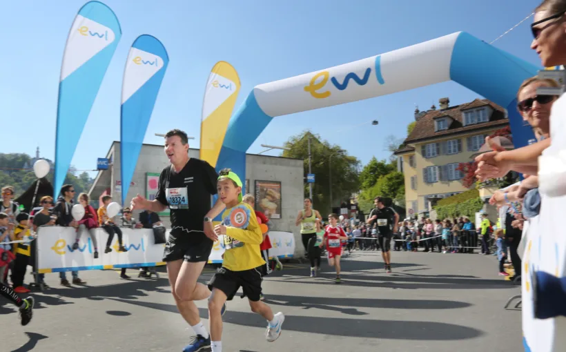 Ein Eventfoto des Luzerner Stadtlaufs am Standort Mühlenplatz. Im Vordergrund sind ein erwachsener und ein jugendlicher Teilnehmer zu sehen. Im Hintergrund sehen wir verschiedene Sponsoring-Elemente von ewl